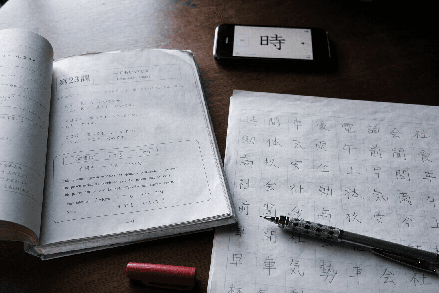 Gros plan d'un bureau d'étude avec manuel Minna no Nihongo ouvert, fiche kanji manuscrite, smartphone avec app japonais, lumière rasante cinématographique